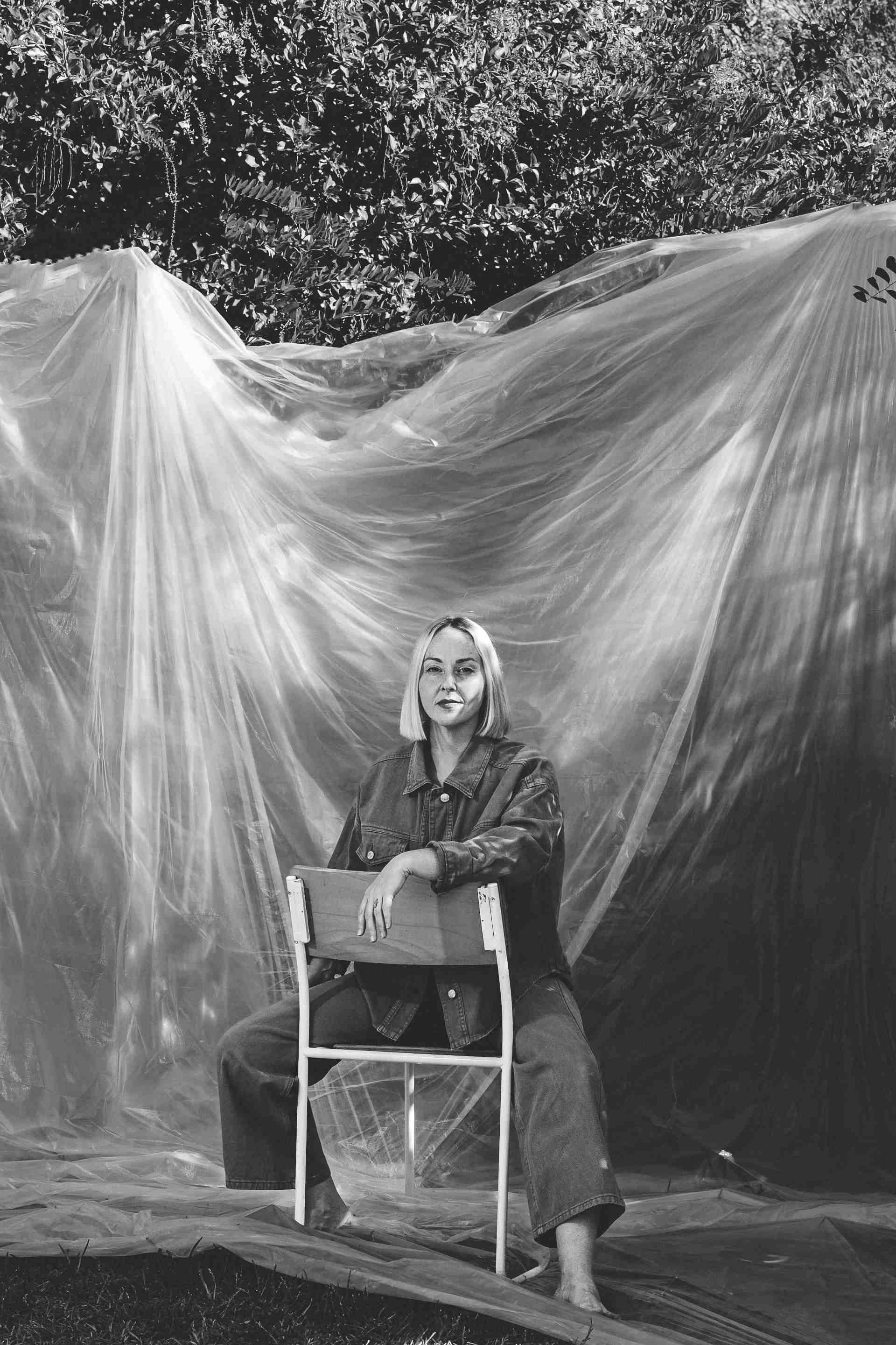Black and white portrait of Anna sitting on a chair, facing the camera, on a large white photo set sheet in a garden setting